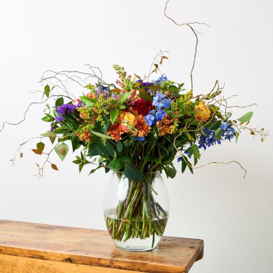 A vibrant multicoloured bouquet with purple, orange, and yellow blooms arranged in a clear glass vase, displayed on a wooden table.