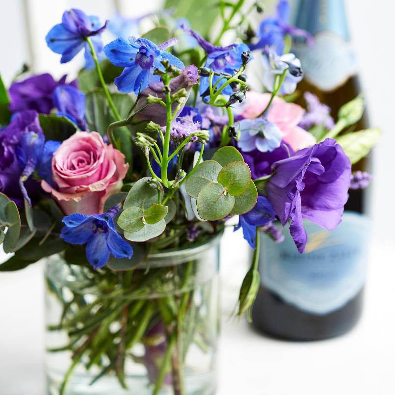A close-up of a vibrant bouquet with blue, purple, and soft pink flowers in a glass vase, paired with a bottle of sparkling wine in the background.