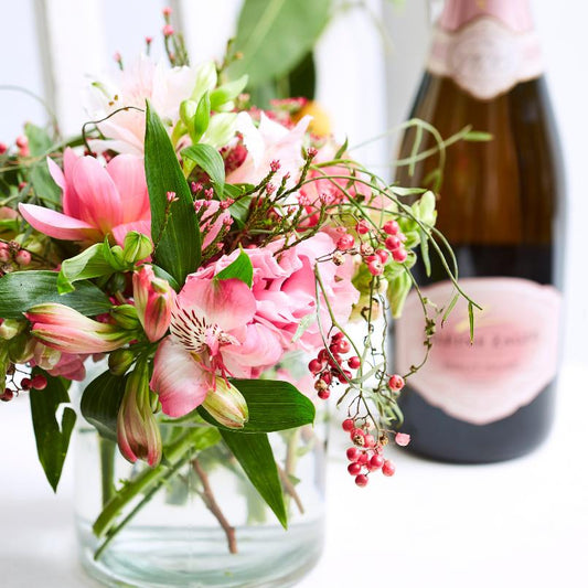 A close-up of a vibrant pink floral arrangement featuring lush pink blooms and greenery in a glass vase, with a bottle of pink sparkling wine in the background.