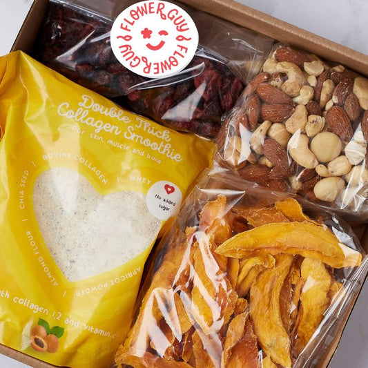 A top-down view of a Flower Guy wellness snack hamper with nutritious treats, including collagen smoothie, dried mango, nuts, and berries.