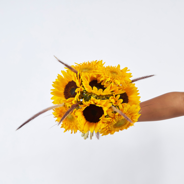 Hand-tied bouquet of sunflowers and golden yellow blooms with dried grass, wrapped in kraft paper, held against a light background