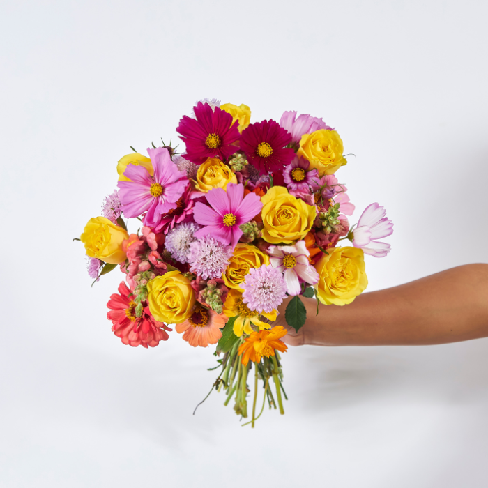 Hand-tied bouquet of bright yellow roses, cosmos, gerberas, and mixed wildflowers, held against a light background.