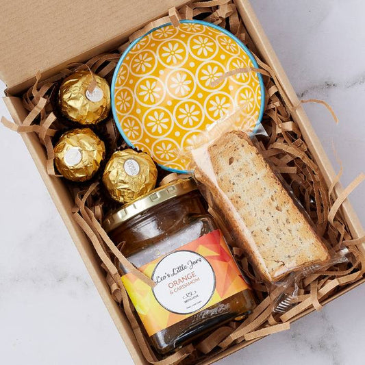 Open gift box with jar of orange marmalade, Ferrero Rocher chocolates, biscotti, and a vibrant yellow ceramic bowl in decorative packaging.