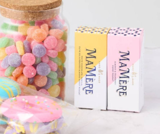 A close-up of the Sweet Harmony Gift Box showing a jar of colourful jelly tots with a cork lid, two Mamère nougat bars in pastel pink and yellow packaging, and a pack of vibrant, frosted biscuits wrapped in plastic.
