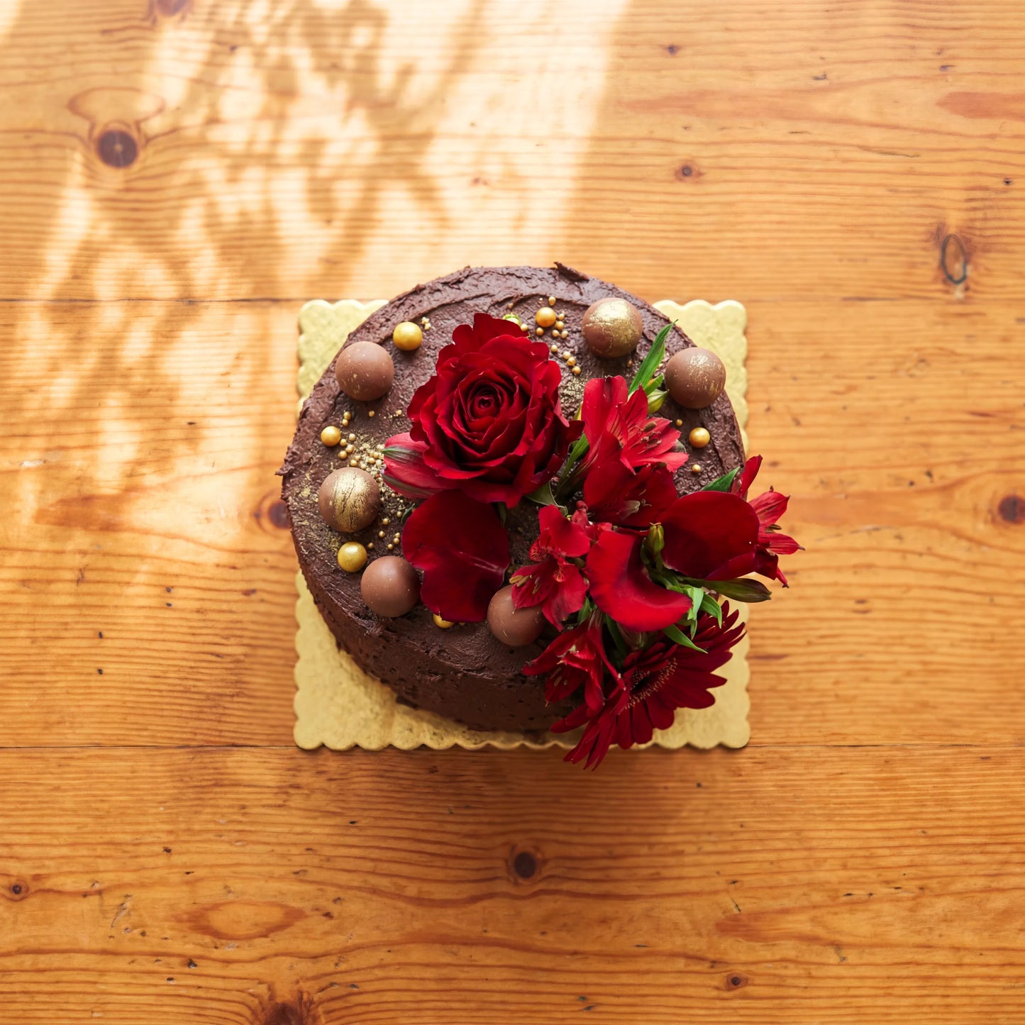 Top-down detail of the Blooming Red Velvet Delight chocolate cake, decorated with milk chocolate truffles, gold-dusted truffles, gold edible pearls and a cluster of fresh red blooms