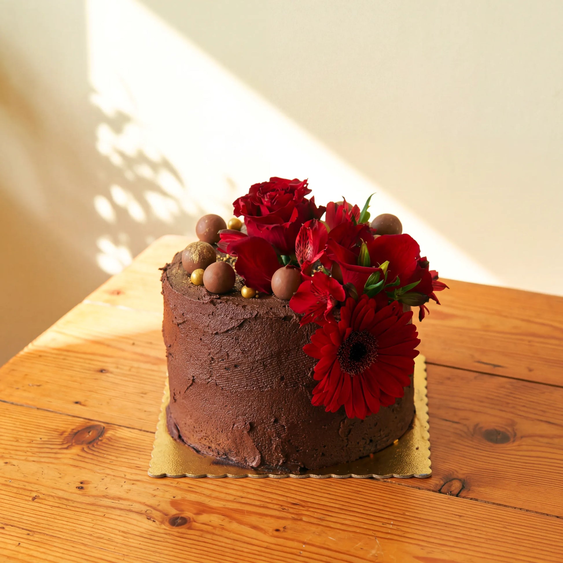 Side angle of the Flower Guy chocolate cake on a gold scalloped board, finished with a fresh red rose and gerbera daisy floral arrangement and chocolate truffle accents