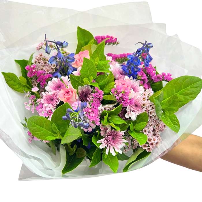 Jump for Joy Bouquet—hand holding pastel mixed flowers in pink and purple with lush greenery, wrapped gift-ready | Flower Guy