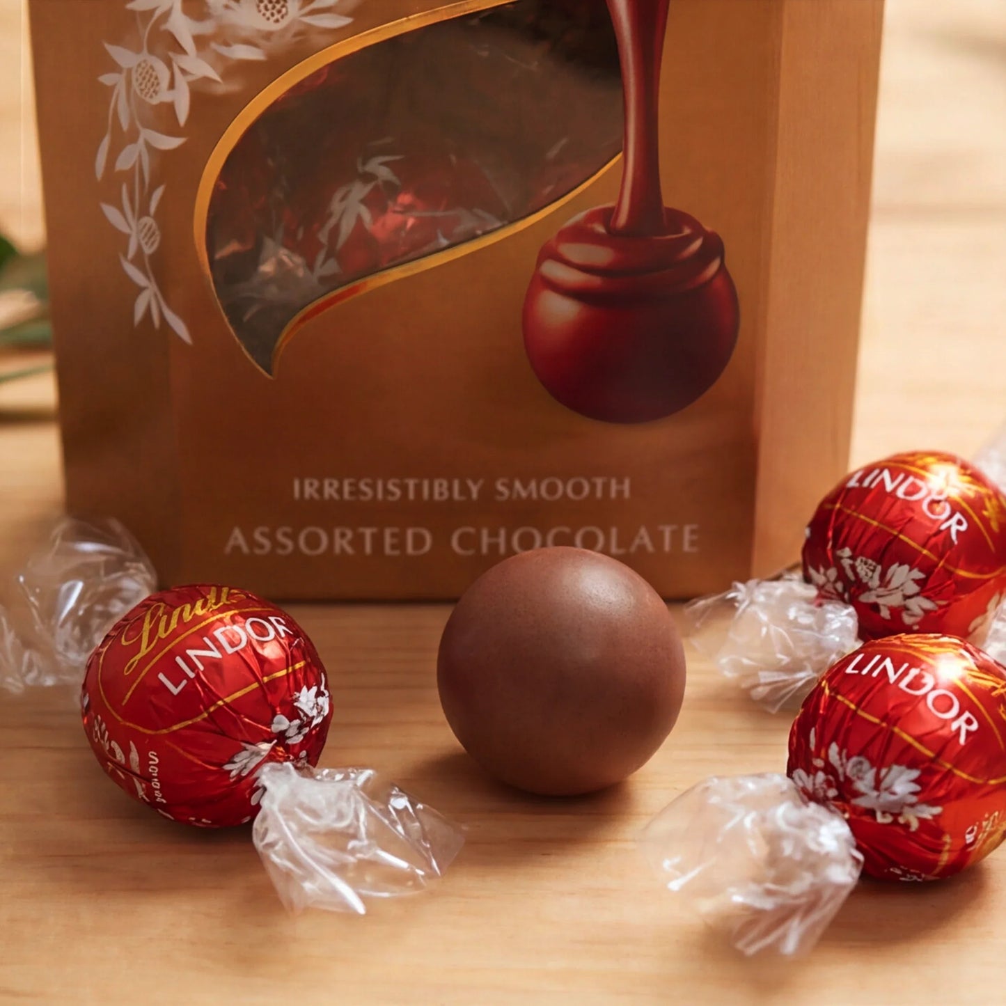 Close-up of Lindt Lindor Assorted Chocolate box with red foil truffles and an unwrapped smooth milk chocolate ball