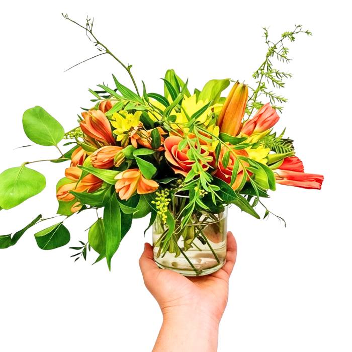Mr Brightside flower arrangement in glass vase with sunlit orange blooms and lush greenery, held in hand | Flower Guy