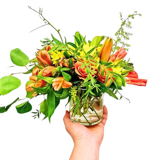 Mr Brightside flower arrangement in glass vase with sunlit orange blooms and lush greenery, held in hand | Flower Guy
