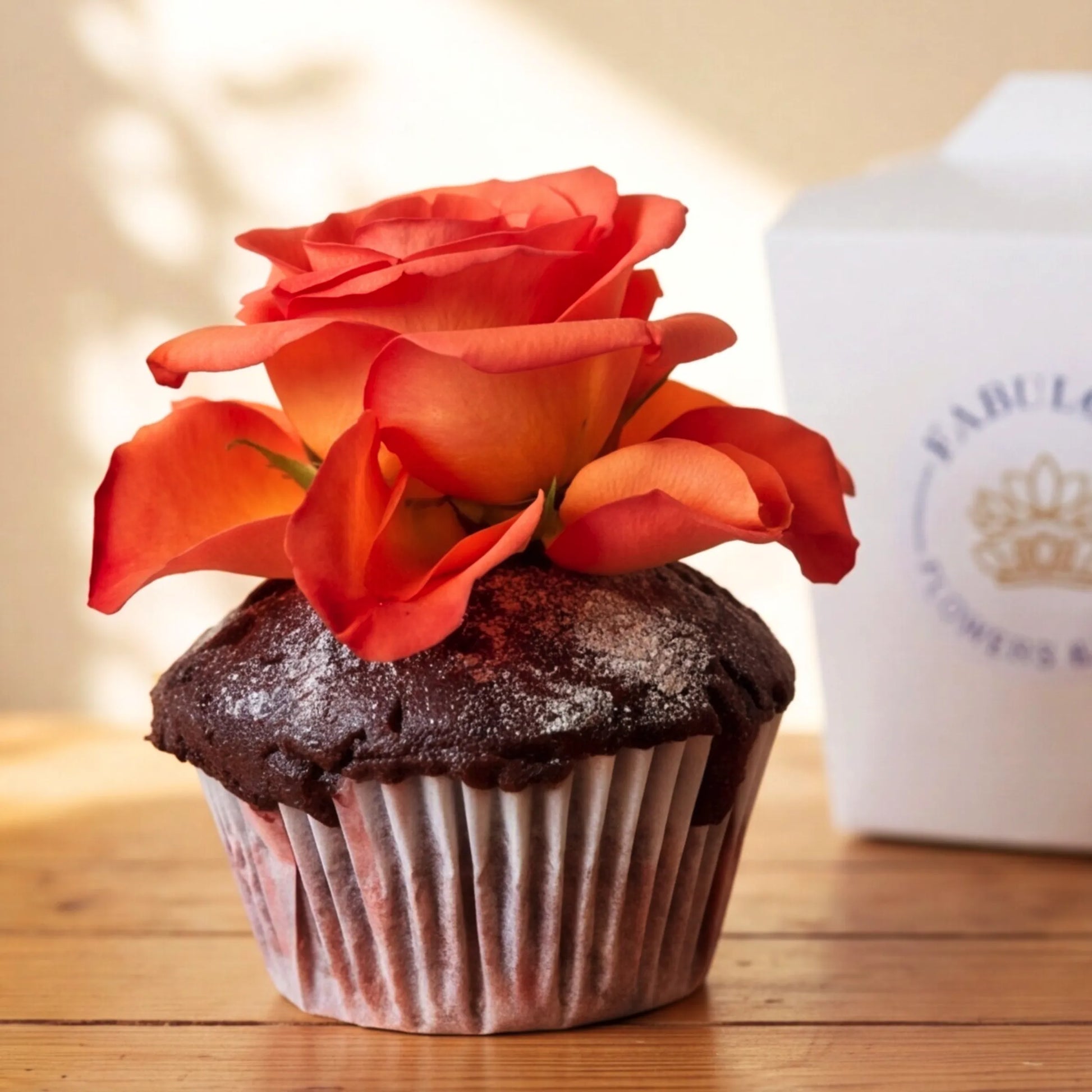 Close-up of a hand-baked chocolate cupcake from Flower Guy with a fresh orange rose topper and a fine icing-sugar dusting on the chocolate dome