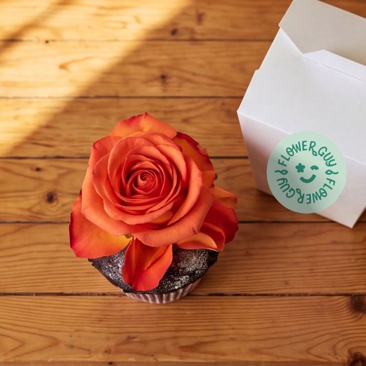 Overhead view of a Flower Guy chocolate cupcake with an orange rose bloom centred on the dark chocolate sponge, white gift box to the side on a rustic wooden surface