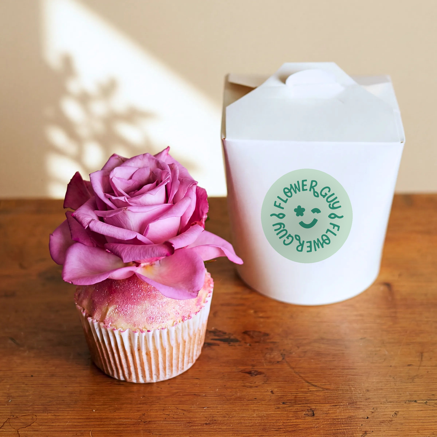 Single Vanilla Cupcake topped with a real pink rose and edible glitter, Flower Guy gift box on a wooden table