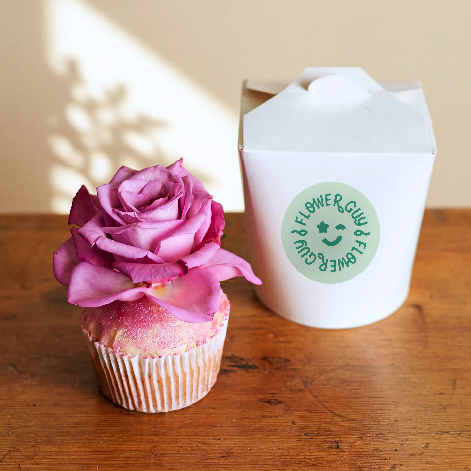 Single Vanilla Cupcake topped with a real pink rose and edible glitter, Flower Guy gift box on a wooden table