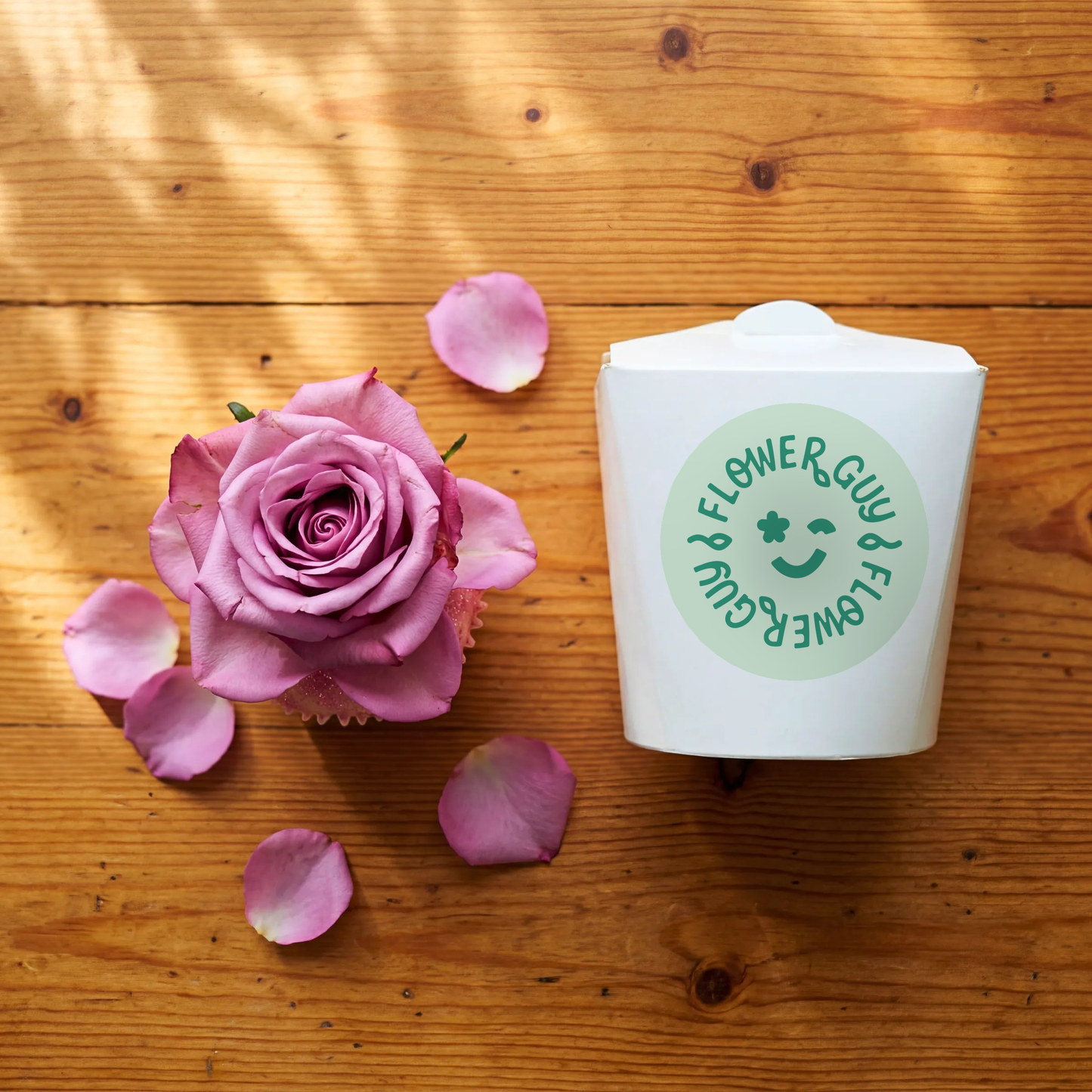 Single Vanilla Cupcake with pink rose, scattered rose petals and Flower Guy gift box on wooden table