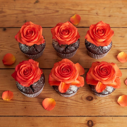 Six chocolate cupcakes arranged in two rows on a rustic wooden table, each topped with a vibrant orange rose and finished with a soft icing-sugar dusting, scattered petals around them