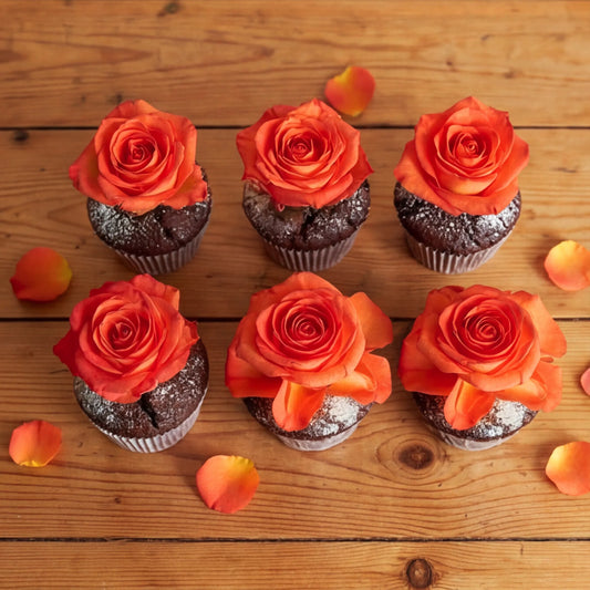 Six chocolate cupcakes arranged in two rows on a rustic wooden table, each topped with a vibrant orange rose and finished with a soft icing-sugar dusting, scattered petals around them