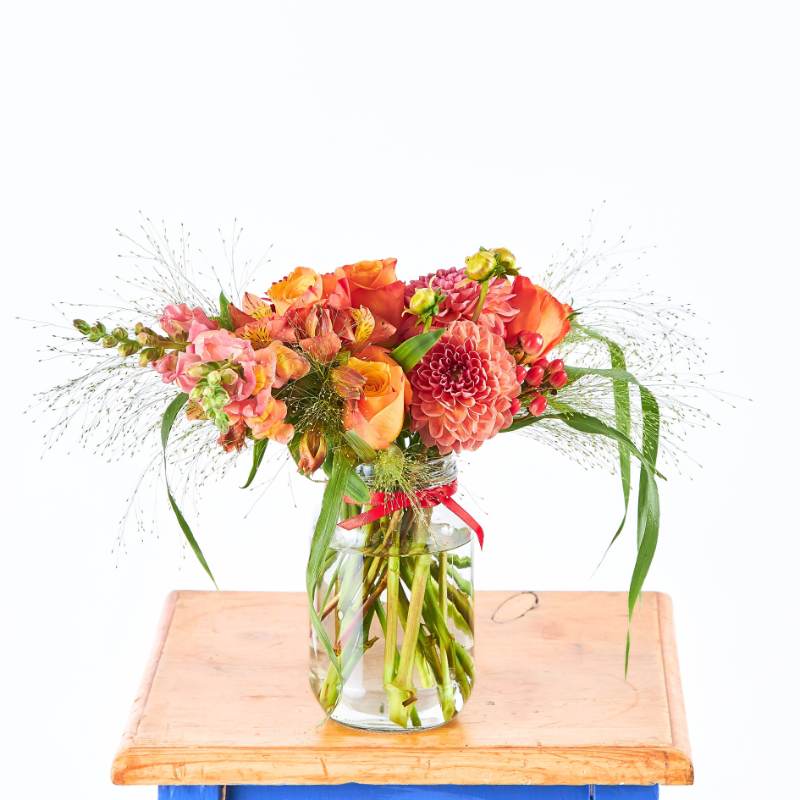Sunset Glow Flower Arrangement in glass jar vase with orange and pink blooms, greenery and berries on wooden board | Flower Guy