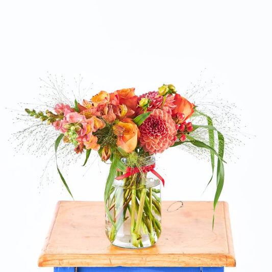 Sunset Glow Flower Arrangement in glass jar vase with orange and pink blooms, greenery and berries on wooden board | Flower Guy