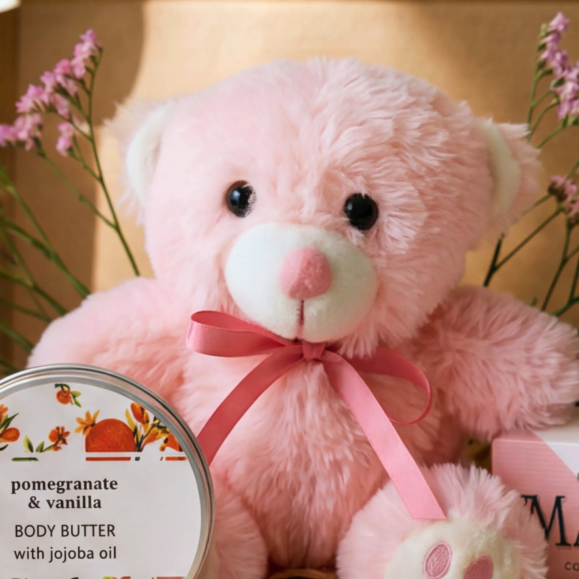 Close-up of pink teddy bear with pink ribbon and pomegranate vanilla body butter in the Teddy Comfort Care Box