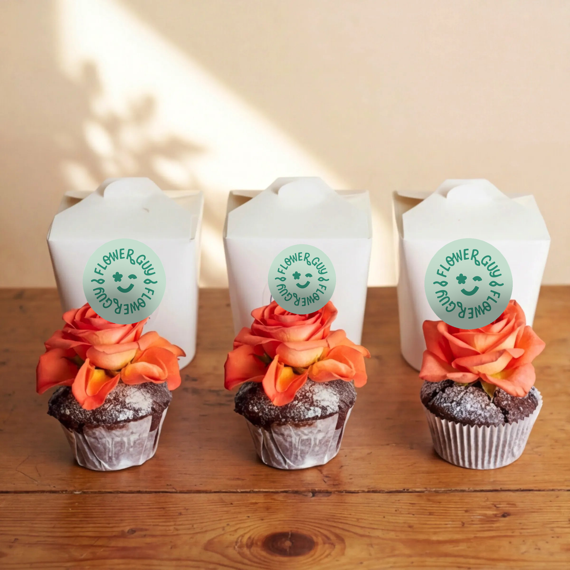 Three chocolate cupcakes lined up on a rustic wooden table in front of three white Flower Guy boxes, each topped with a vibrant orange rose and finished with a soft icing-sugar dusting