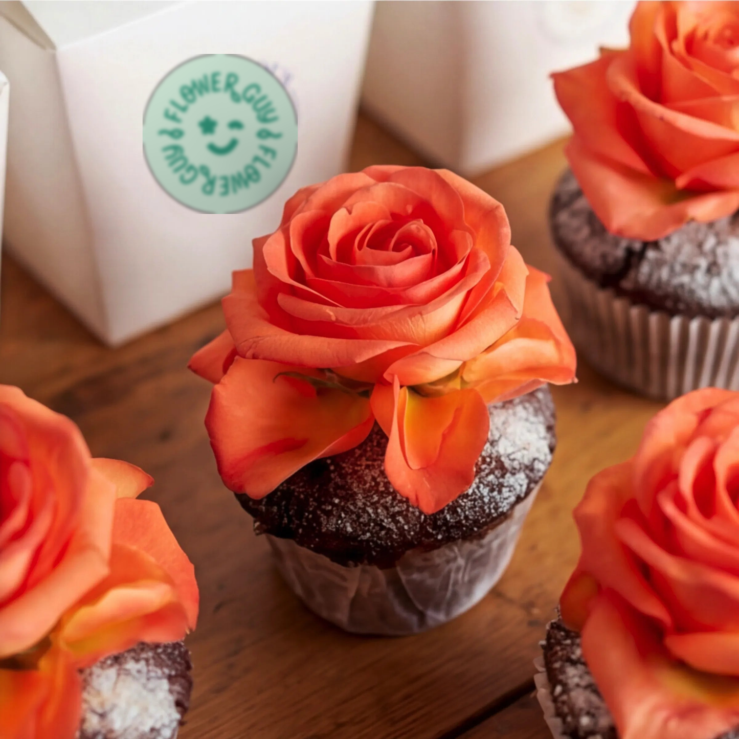 Close-up of Flower Guy chocolate cupcakes with vibrant orange rose toppers and icing-sugar finish on dark chocolate sponge, alongside the white Flower Guy gift box
