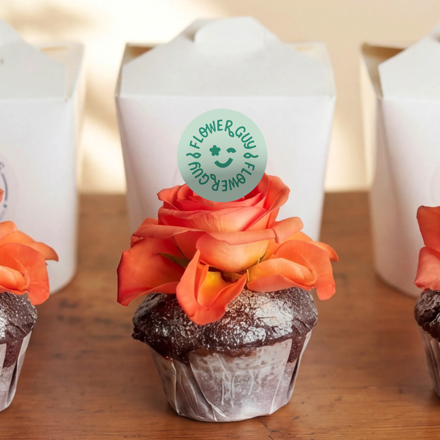 Macro of a single chocolate cupcake from the Three Chocolate Cupcakes set, dark chocolate sponge with icing-sugar dusting and an open orange rose bloom resting on top