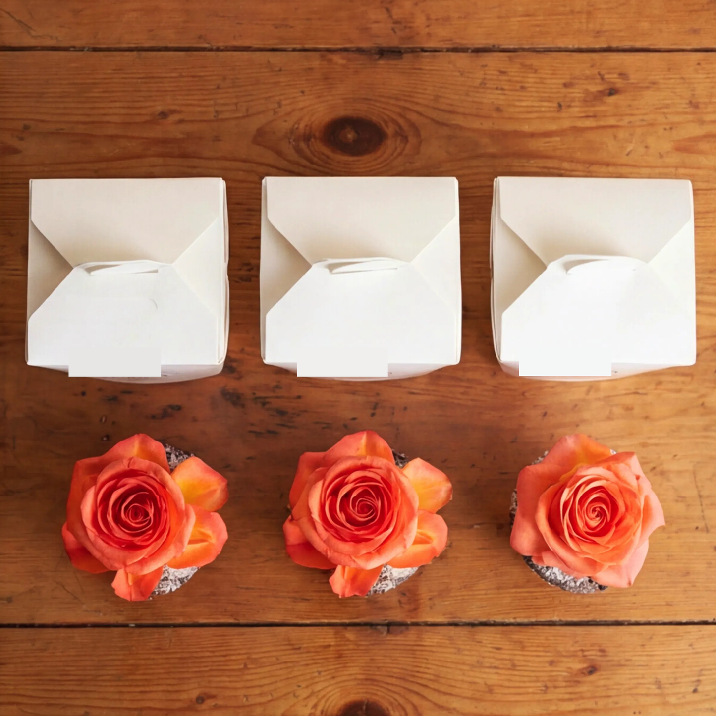 Overhead flat lay of three white Flower Guy cupcake boxes paired with three orange rose-topped chocolate cupcakes arranged on a rustic wooden surface