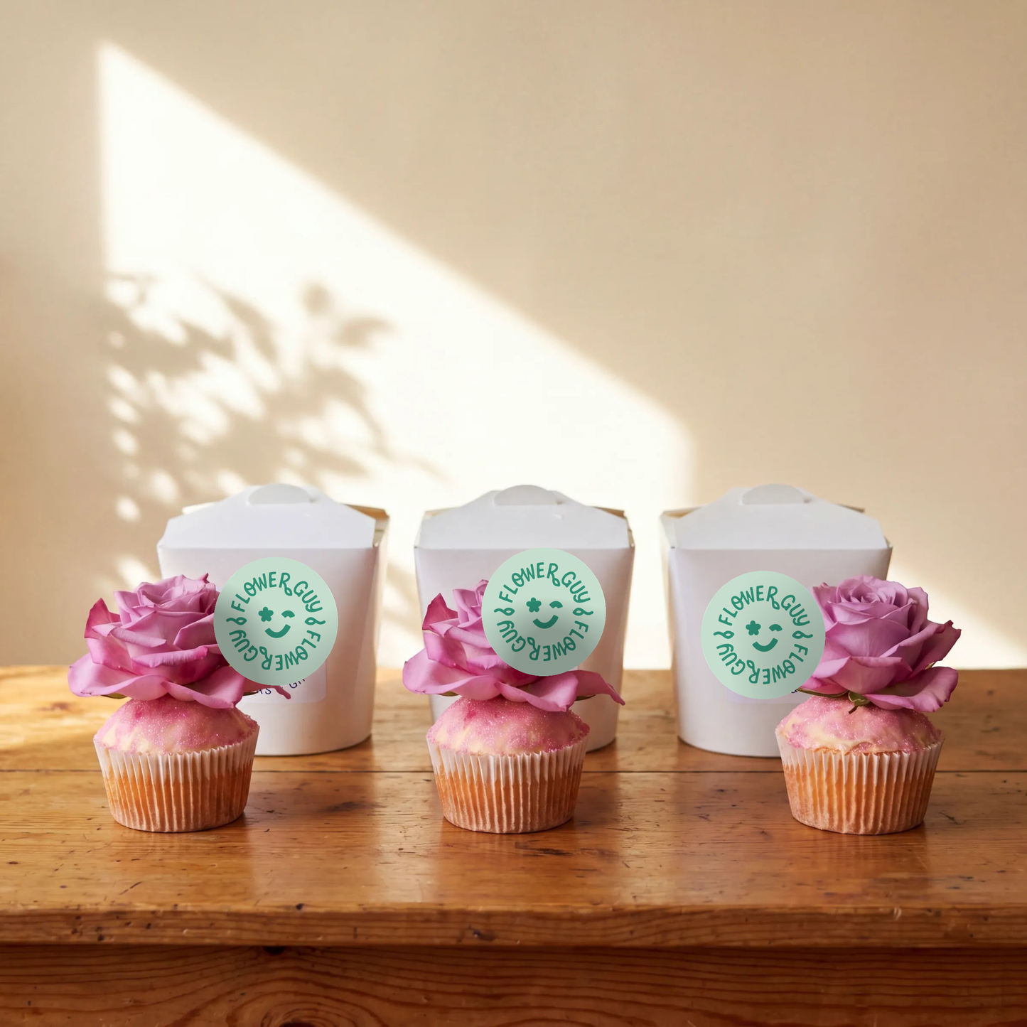 Three vanilla cupcakes lined up on a rustic wooden table in front of three white Flower Guy boxes, each cupcake topped with a fresh pink rose and pink edible glitter