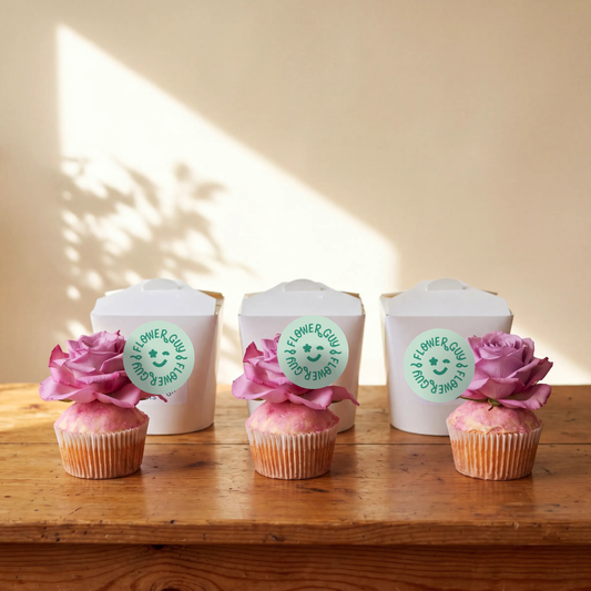 Three vanilla cupcakes lined up on a rustic wooden table in front of three white Flower Guy boxes, each cupcake topped with a fresh pink rose and pink edible glitter