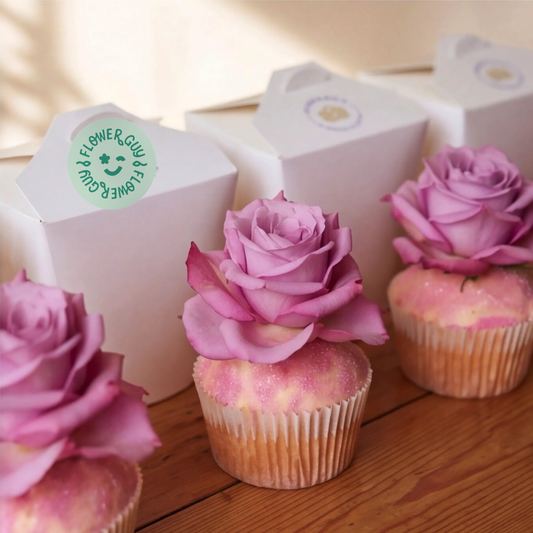 Close-up of three vanilla cupcakes with pink rose toppers and edible glitter, paired with white Flower Guy gift boxes